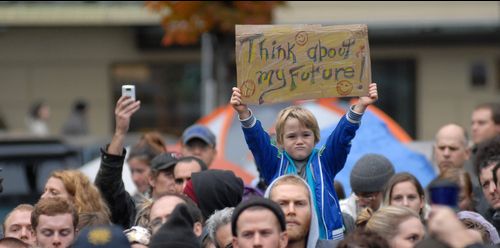 Photo of protestors.  A small boy is sitting on someone's shoulders. He is holding up a sign saying "Think about my future!"